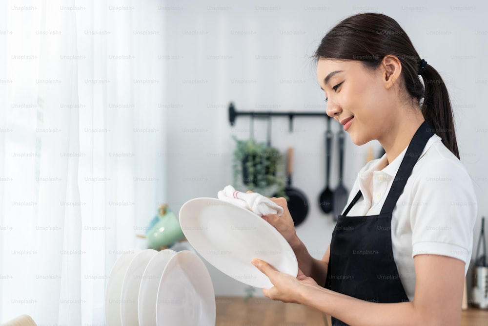 Servizio di pulizia attivo asiatico donna che pulisce in cucina a casa. La bella ragazza delle pulizie si sente felice e pulisce i piatti dopo il lavaggio per le faccende domestiche o le faccende domestiche.