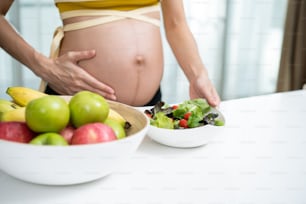 Close up of Asian woman pregnant standing and holding mix slad bowl. Attractive beautiful young female pregnancy stand and touching baby in the womb, choose healthy food or vegetarian food in kitchen.