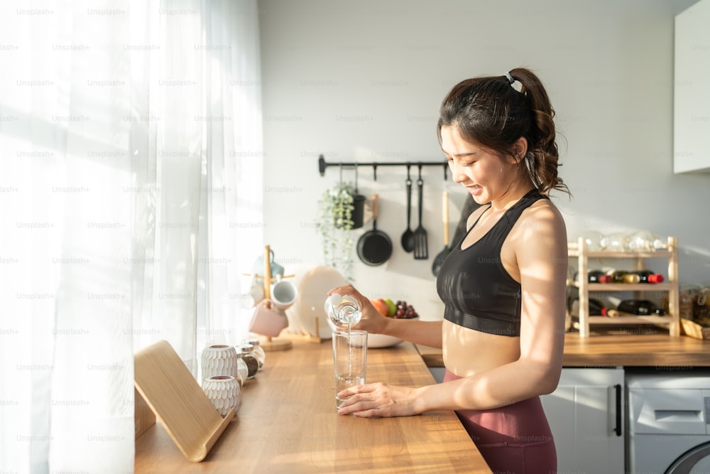 Asian beautiful woman in sportswear drink water after exercise at home. Young thirsty active sport girl takes a sips of clean mineral natural in cup after workout for health care in kitchen in house.