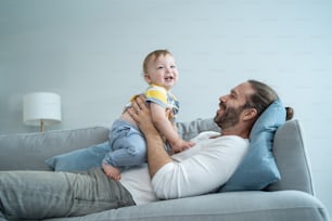 Caucasian loving dad play with baby boy child on sofa in living room. Happy family, attractive handsome young father lifting adorable toddler son fly up enjoy doing airplane game in morning in house.