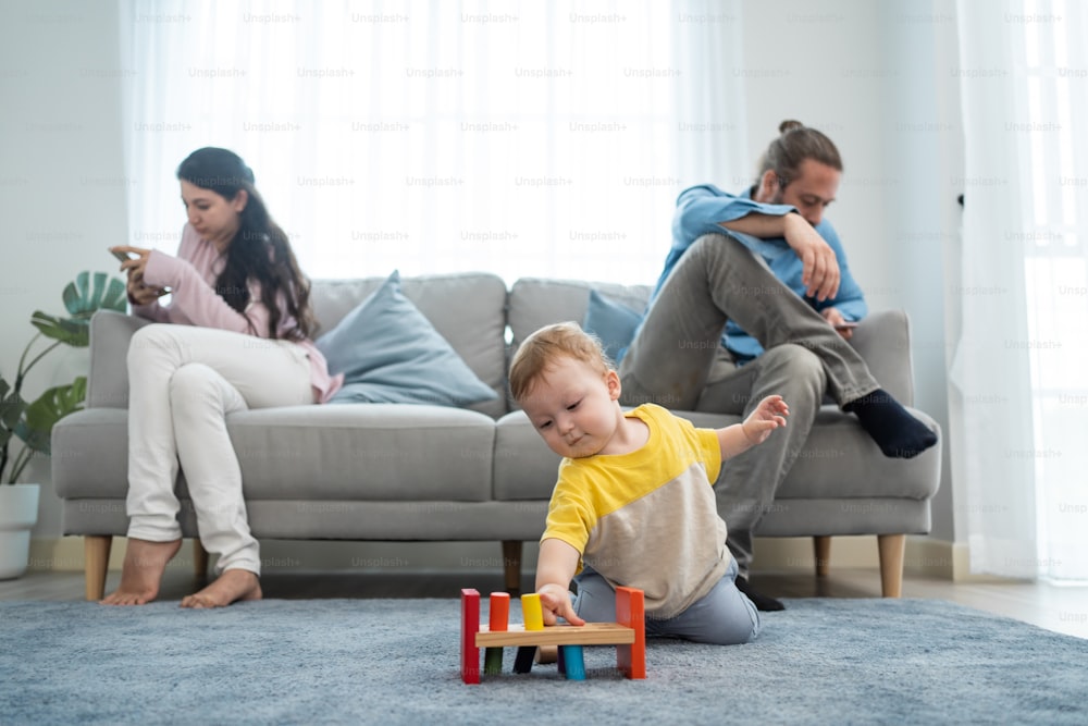 Caucasian phone addict parents don't pay attention with baby toddler. Family problem, Father and Mother sit on sofa and using cellphone ignore young infant boy son play alone on floor in living room.
