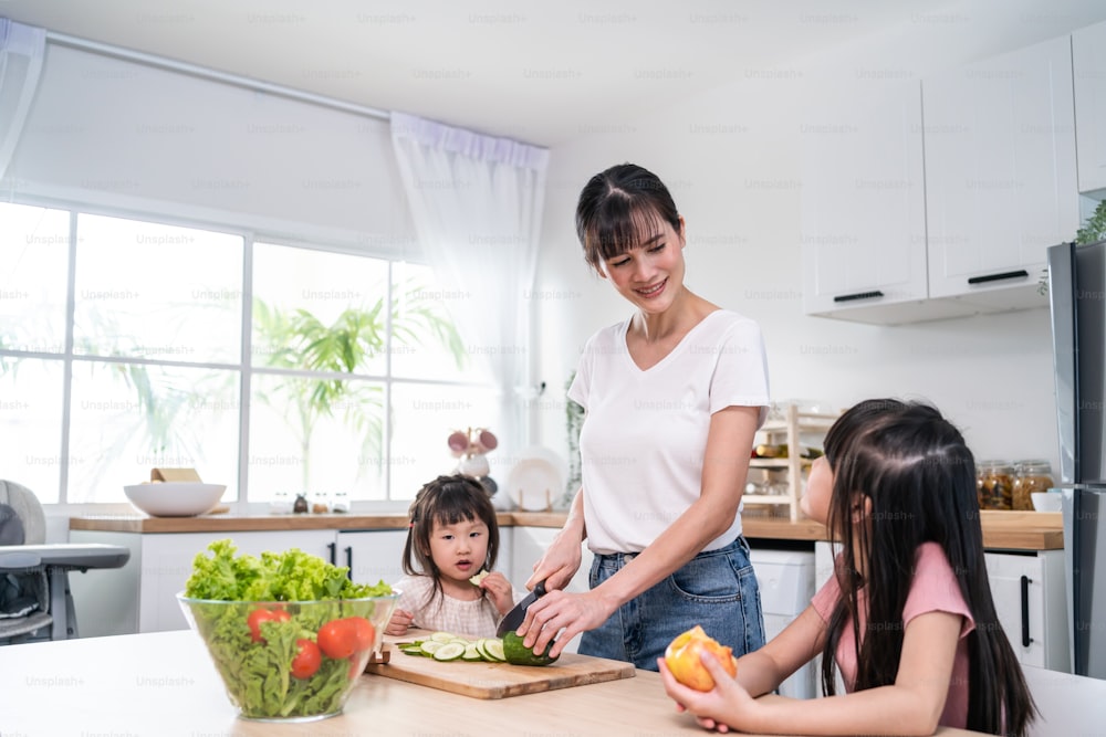 Asian beautiful mother cooking vegetable salad with two young daughter. Happy Family, attractive caring parent mom enjoy preparing healthy foods for little girl child sibling sister in kitchen at home