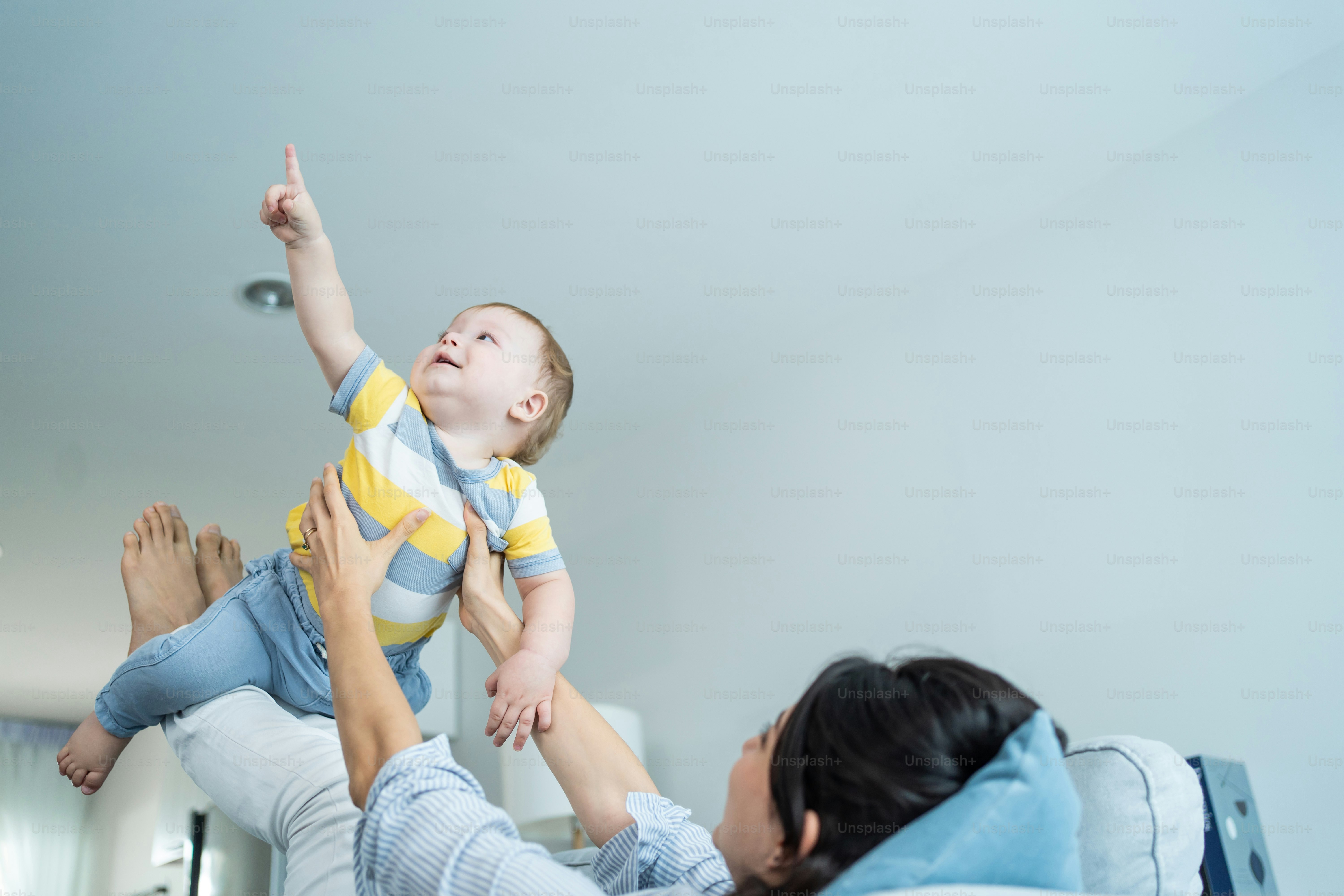 Caucasian loving mom play with baby boy child on sofa in living room. Happy family, attractive beautiful caring young mother lifting toddler son fly up enjoy doing airplane game in morning in house.