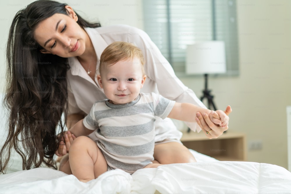 Caucasian loving mom play with cute baby boy child on bed in bedroom. Happy family, attractive beautiful caring young mother hugging toddler kid son enjoying activity relationship in morning in house.