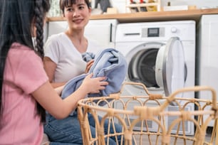 Asian happy family stay home, mother teach daughter wash dirty clothes. Young little cute girl child help and learn from parent mom to put laundry in washer appliance at home. Domestic-Housekeeping.