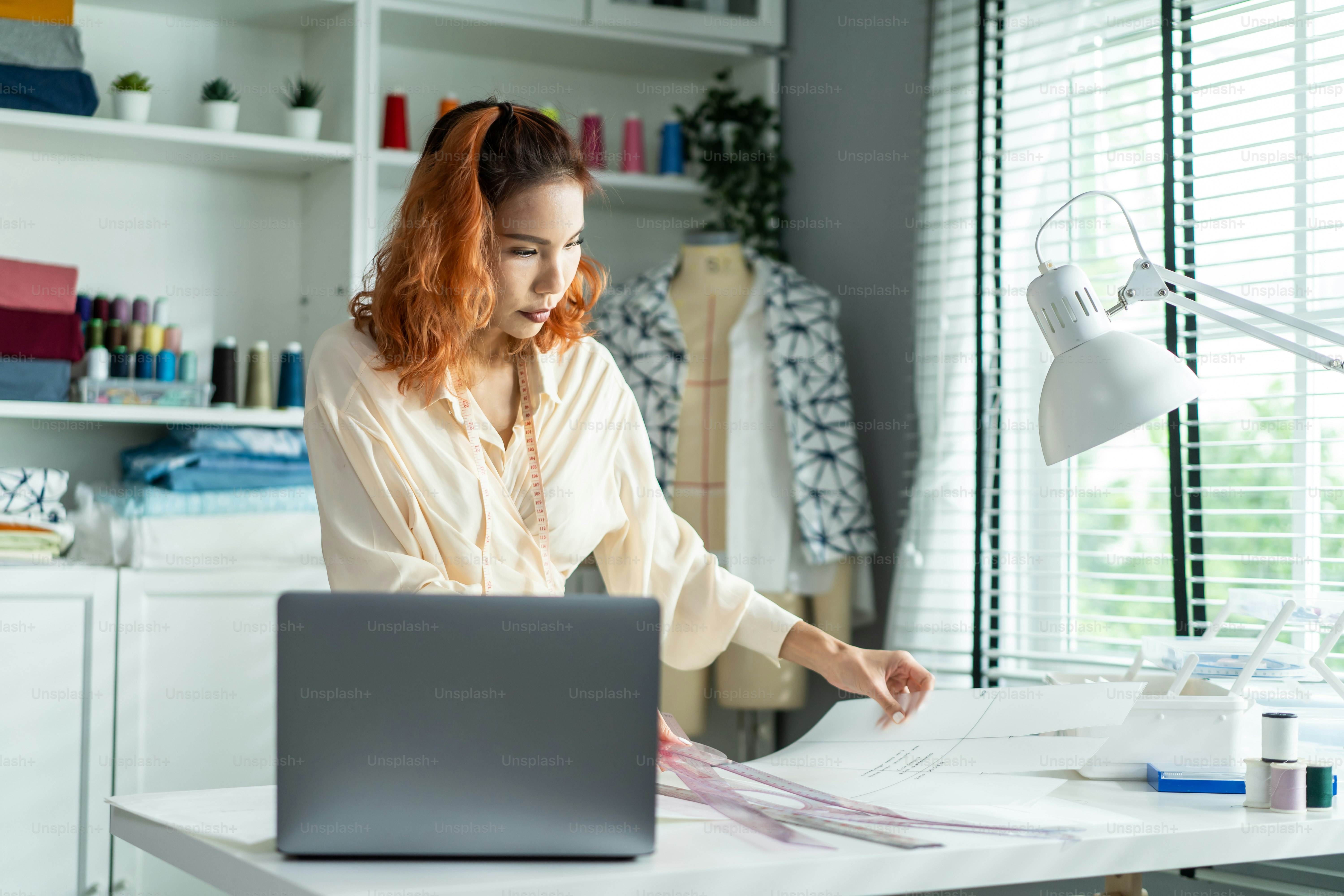 Asian tailor woman working to design new clothes in tailoring atelier. Attractive young female fashion designer dressmaker create new pieces of clothing from patterns and designs in workshop room.
