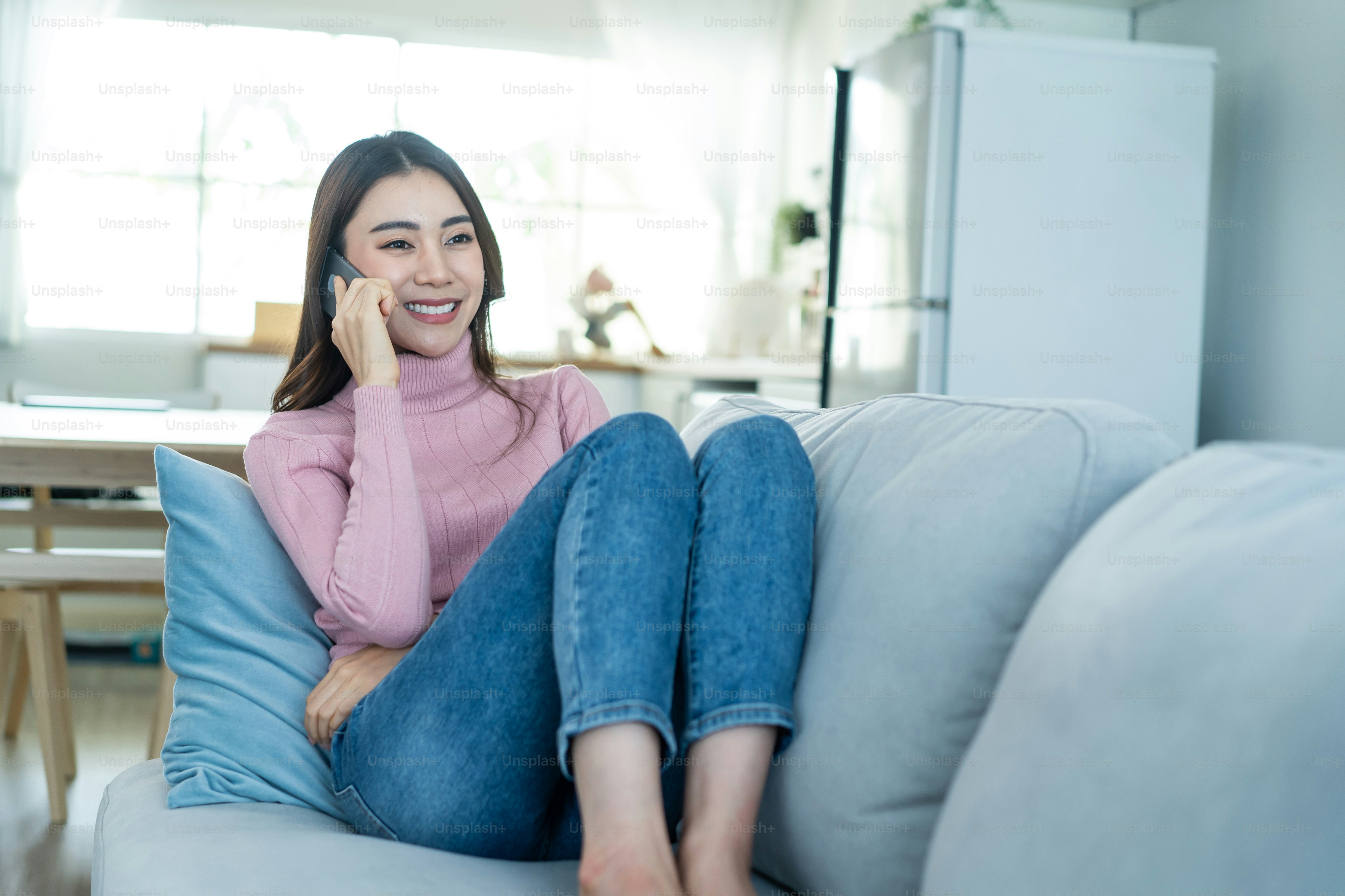 La bella donna asiatica si siede sul divano e parla al telefono cellulare in casa. La giovane ragazza attraente felice trascorre il tempo libero a casa, si rilassa e si diverte a comunicare e discutere sullo smartphone in salotto.
