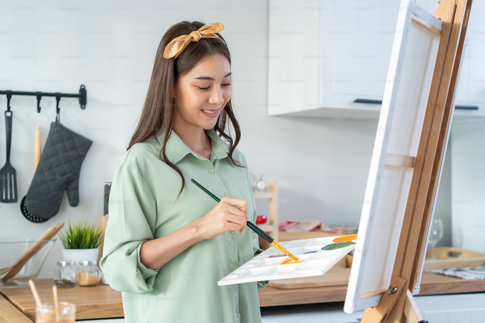 Asian young talented woman artist coloring on painting board in house. Attractive beautiful female draw art picture, creating artwork with watercolor paint and brush enjoy creativity activity at home.
