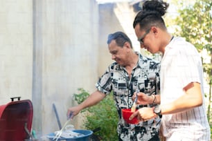 Older man preparing meat on a barbecue grill for his family's lunch in the backyard. His son is standing next to him.