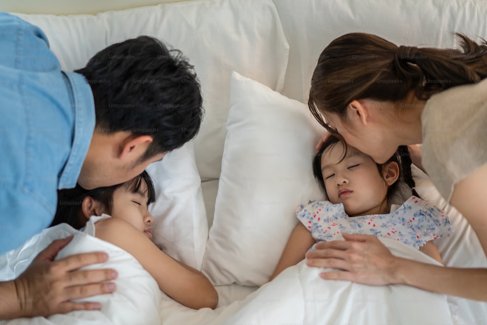 Asian loving parents take care of sleeping daughter in bedroom at home. Attractive happy family couple, mother and father puts blanket on comfortable asleep napping little baby kid on bed in morning.