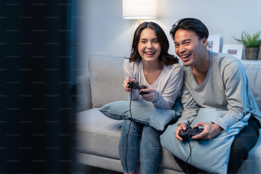 Asian young man and woman couple enjoy playing game together in house. Attractive funny male and female friend feeling happy sit on sofa in living room, spend time together play joystick game at home.