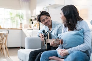 アジアの若い愛するカップルは、自宅のリビングルームで一緒にギターを弾きます。魅力的なロマンチックな新婚、ソファに座ってアクティブなロマンチックな男性と美しいガールフレンドが自宅で楽器を演奏することを教えます。