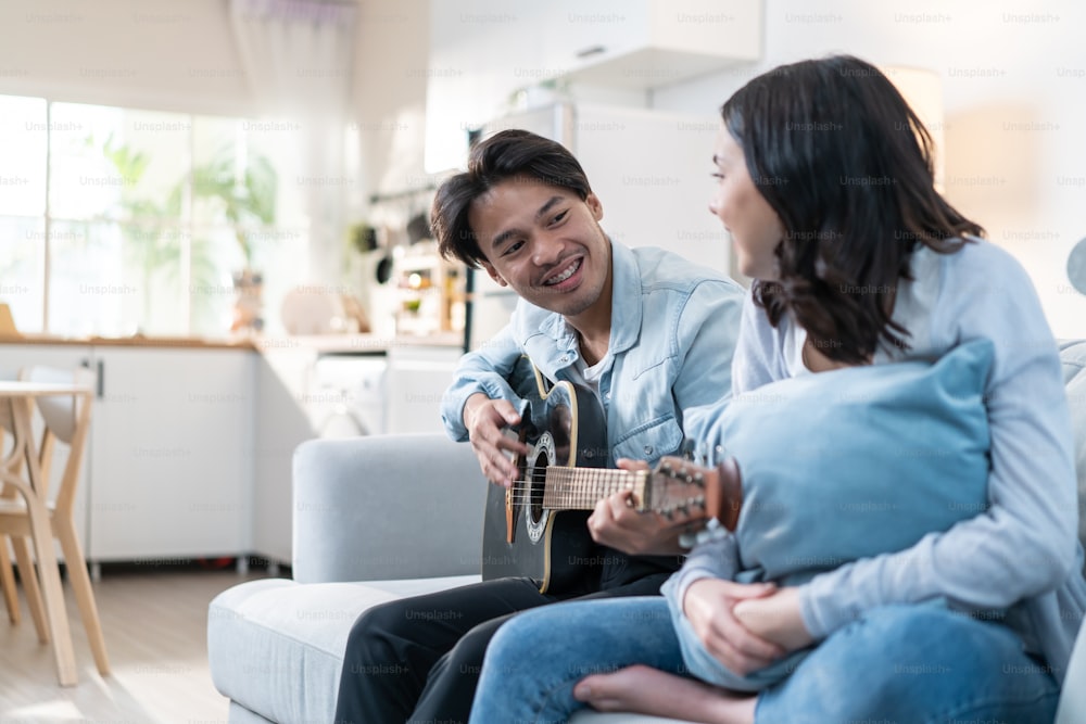 Asian young loving couple play guitar together in living room at home. Attractive romantic new marriage, active romantic man sitting on sofa and teach beautiful girlfriend play an instrument at home.