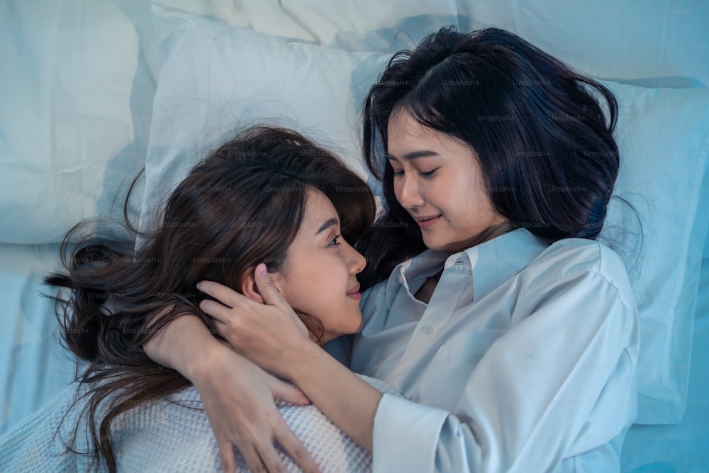 Asian beautiful lesbian couple lying down on bed and hugging each other. Attractive romantic girl friend in pajamas spending night leisure time together in bedroom. Homosexual freedom pride concept.