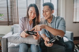 Happy Asian couple having fun playing video game, man teaching woman how to play game at home