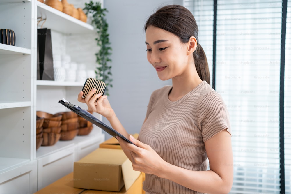 Asian beautiful woman check vase goods order for customer from shelf. Young attractive business girl working to preparing parcel boxes checking ecommerce shipping online retail to sell at home store.