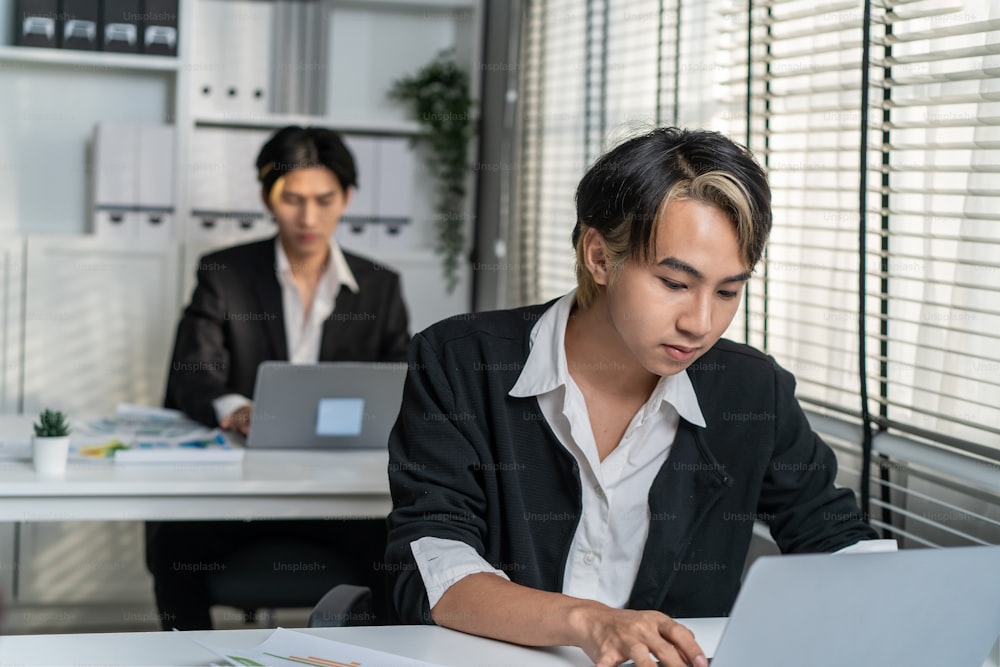 Asian handsome businessman gay people working in office with happiness. Attractive male lgbtq collegues in formal wear sitting on table, use laptop computer with gay pride and confidence at workplace.