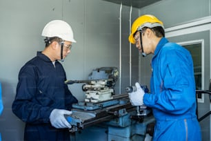 Trabalhadores mecânicos asiáticos que trabalham na fresadora. Os técnicos usam óculos de proteção e capacete ao operar a máquina por precaução de segurança. Líder aconselhando o membro de sua equipe a fazer um trabalho