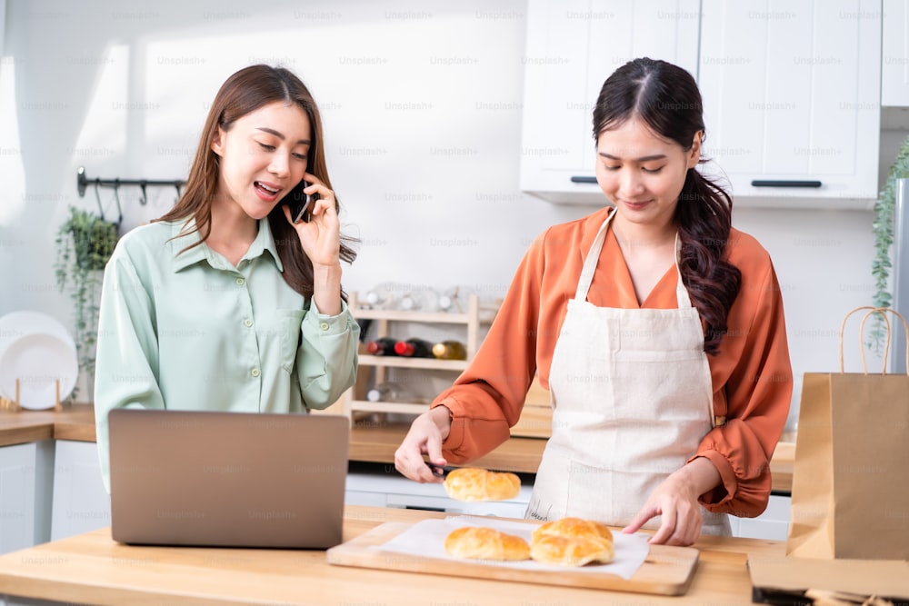 Asian beautiful business woman and friend bake bakery and sale in house. Attractive lesbian couple feel happy and exciting for success in entrepreneur sell food order from home then high five together