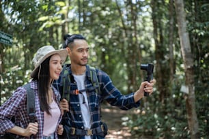 Asian young couple backpacker vlogger traveling in the forest alone. Attractive man and woman traveler use camera record video vlog, walking in nature wood with happiness during holiday vacation trip.