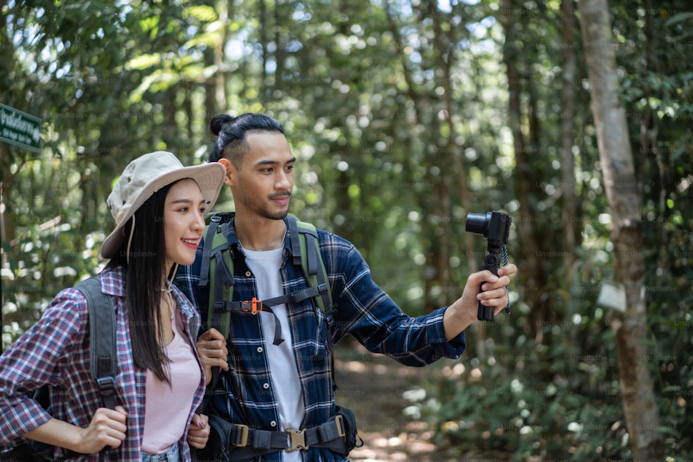 Asian young couple backpacker vlogger traveling in the forest alone. Attractive man and woman traveler use camera record video vlog, walking in nature wood with happiness during holiday vacation trip.