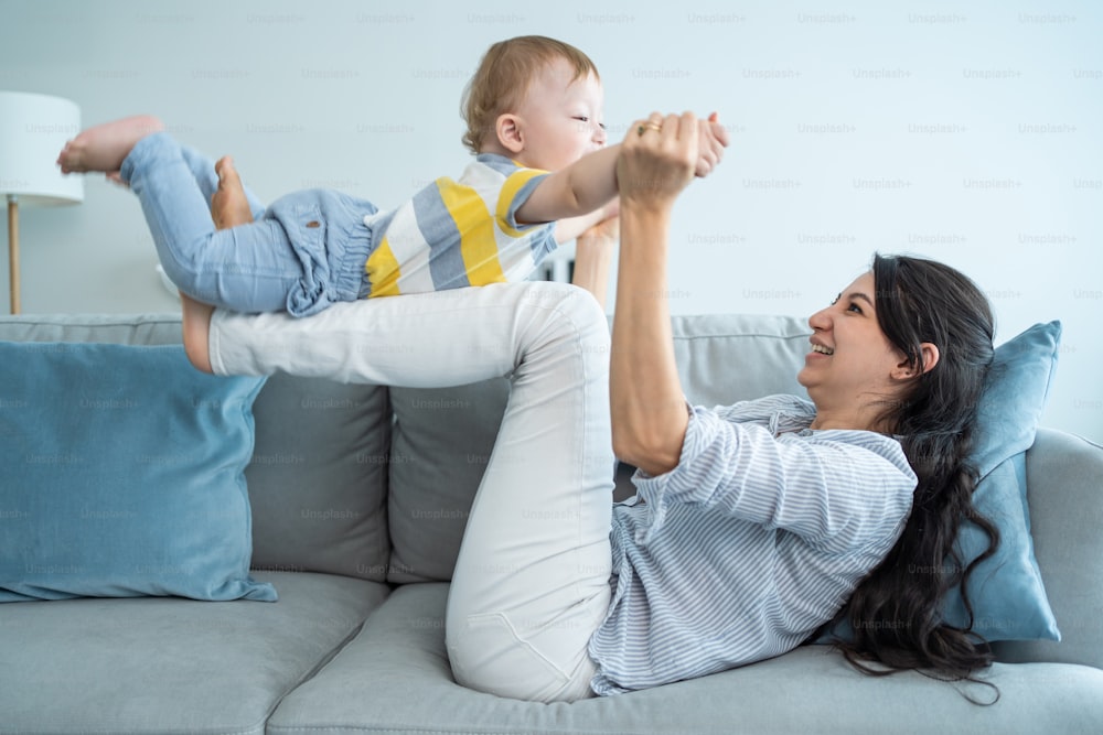 Caucasian loving mom play with baby boy child on sofa in living room. Happy family, attractive beautiful caring young mother lifting toddler son fly up enjoy doing airplane game in morning in house.