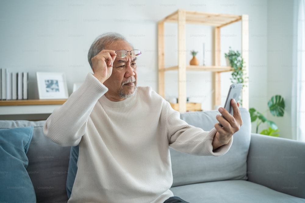 Asian senior elderly male using mobile phone in living room at home. Strong older mature grandfather wear eyeglasses, sit on sofa and chat on smartphone communicate with family enjoy retirement life.