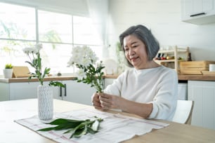 Asian senior old woman puts flowers on vase with happiness in kitchen. Beautiful active elderly grandmother sit on table, feeling happy while decorate and renovate apartment. Activity at home concept.
