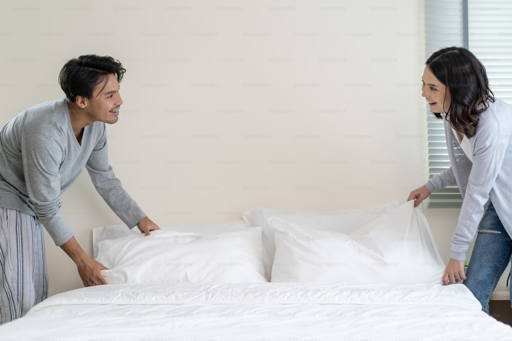 Asian young couple folding blanket after wake up together in morning. Attractive new marriage man and woman in pajamas feel happy and relax in early after get up from bed in bedroom together at home.