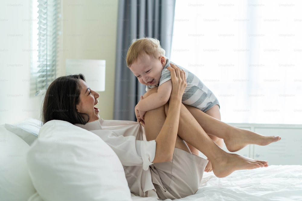 Caucasian loving mom play with cute baby boy child on bed in bedroom. Happy family, attractive beautiful caring young mother lifting toddler son fly up enjoy doing airplane game in morning in house.
