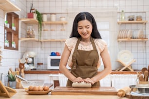 Mujer madura asiática hermosa que se queda en casa, pasa tiempo cocinando en la cocina. Joven atractiva que lleva el delantal y la sonrisa, masa de levadura amasada con la mano para hornear la panadería en la mesa para la cena en casa