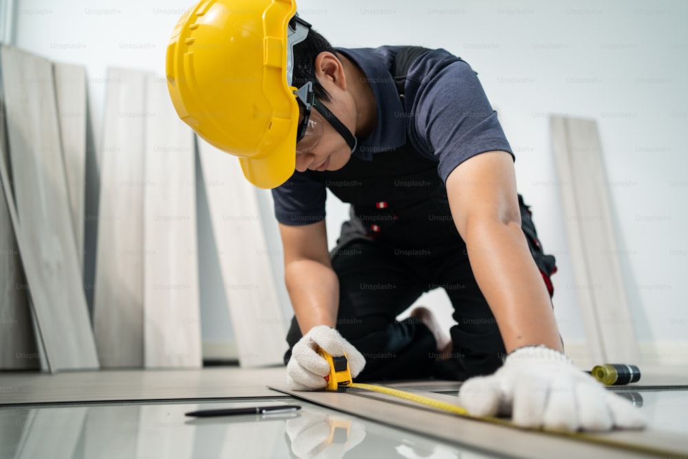 アジアの職人や大工の労働者の男性は、作業用安全ヘルメットと眼鏡を着用し、廊下で巻尺の寄木細工の床を使用します。ジョイナービルダーの男性は、アパートや家を改装するために床にラミネートボードを設置します