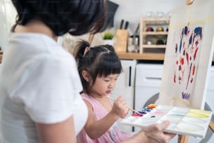 Asiatische junge glückliche Kind-Tochter malen auf Malbrett mit Mutter. Glückliche Familienaktivität, kleines Mädchen lernen, wie man Kunstbild mit Aquarellfarbe und Pinsel zeichnet und Kreativität mit Mama genießt.