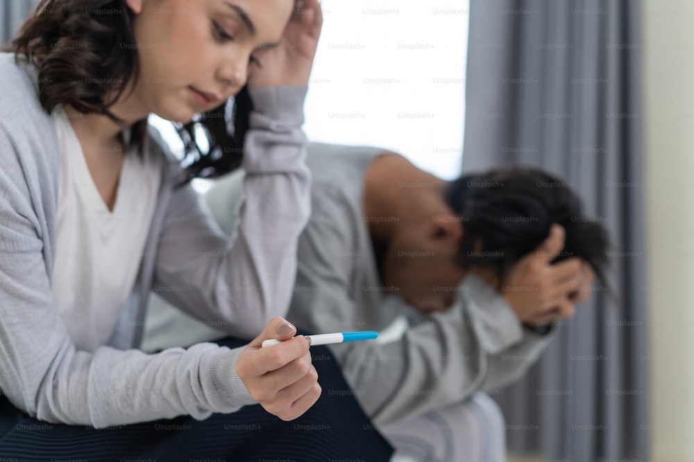 Asian young couple feeling shock after look at positive Pregnancy test. Disappointed unplanned pregnancy men and women sit on bed in bedroom with upset and sad face for problem don't want to pregnant.