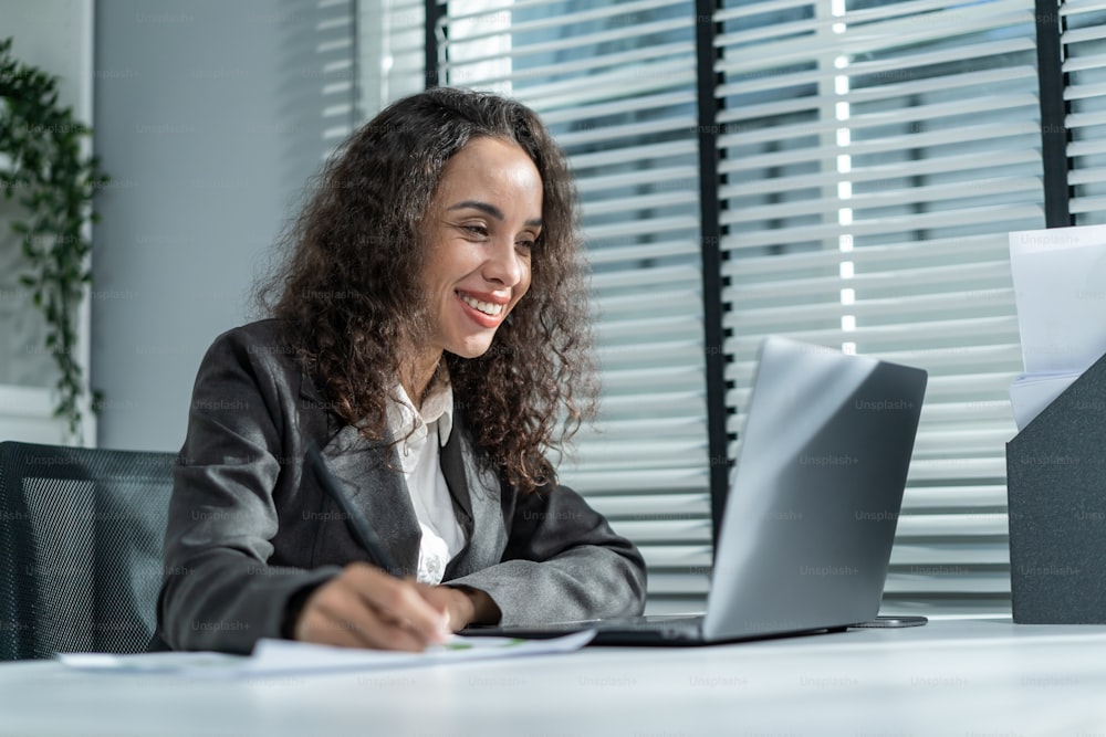 Happy beautiful Latino businesswoman in formal suit work in workplace. Attractive female employee office worker smile, use laptop computer for discuss with co worker and take note on paper in company.