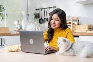 아시아의 매력적인 사업가가 노트북에 타이핑하고 고양이를 본다. 젊고 아름다운 여성 직장인은 팬데믹 기간 동안 집에서 일하는 동안 테이블에 앉아 행복을 느끼고 애완 동물을 키우는 것을 즐깁니다.