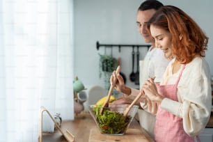 Asian beautiful woman cook green salad with husband in kitchen at home. Young attractive couple feeling happy and enjoy eating vegetables healthy foods to diet and lose weight for health care in house