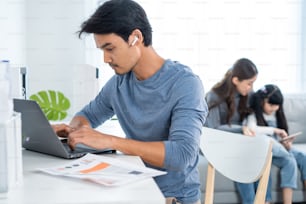 Asian business man father sit on table work at home with family behind. Male freelance worker using laptop computer talk to colleagues on virtual video call conference online in living room at house.