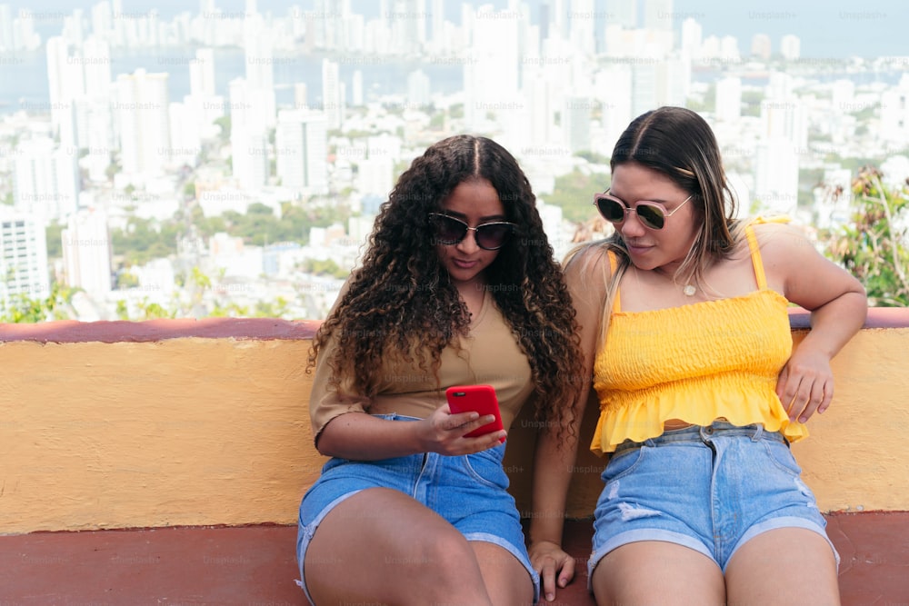 Zwei Latina-Frauen sitzen auf der Straße der Stadt, schauen aufs Handy und unterhalten sich auf der Straße.
