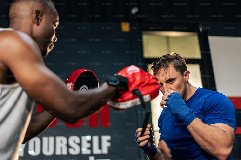Hombre caucásico usa guantes de boxeo golpeando adelante con entrenador africano. Atractivo joven culturista boxeador masculino que se ejercita con la ayuda del entrenador afroamericano para el cuidado de la salud en el gimnasio o club de fitness.