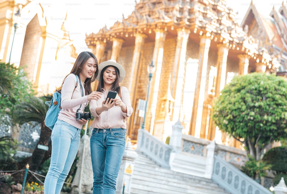 Mulheres asiáticas turísticas amigas usando o telefone inteligente e desfrutar de passeios enquanto viajam no templo do buda esmeralda, Wat Phra Kaew, lugar turístico popular em Banguecoque, Tailândia