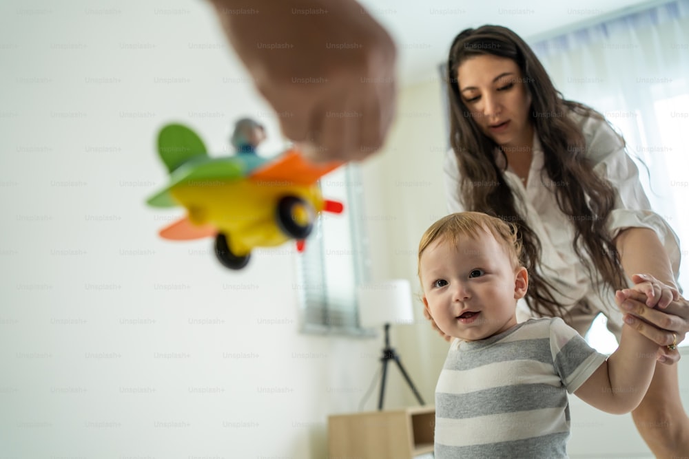 Caucasian baby boy child learn to walk with parents support in house. Happy family, mother and father helping young toddler son taking first step walk on floor to develop skill in living room at home.