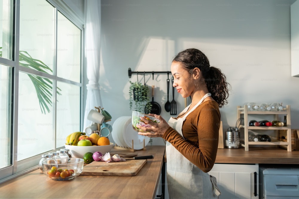 Latino attractive woman wear apron cook green salad in kitchen at home. Young beautiful girl feeling happy and enjoy eating vegetables healthy foods to diet and lose weight for health care in house.