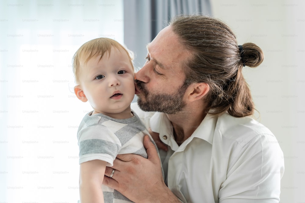 Caucasian loving father kiss on baby boy child cheek in living room. Happy family, attractive young caring daddy holding his toddler son on hands with happiness in house. Activity relationship at home