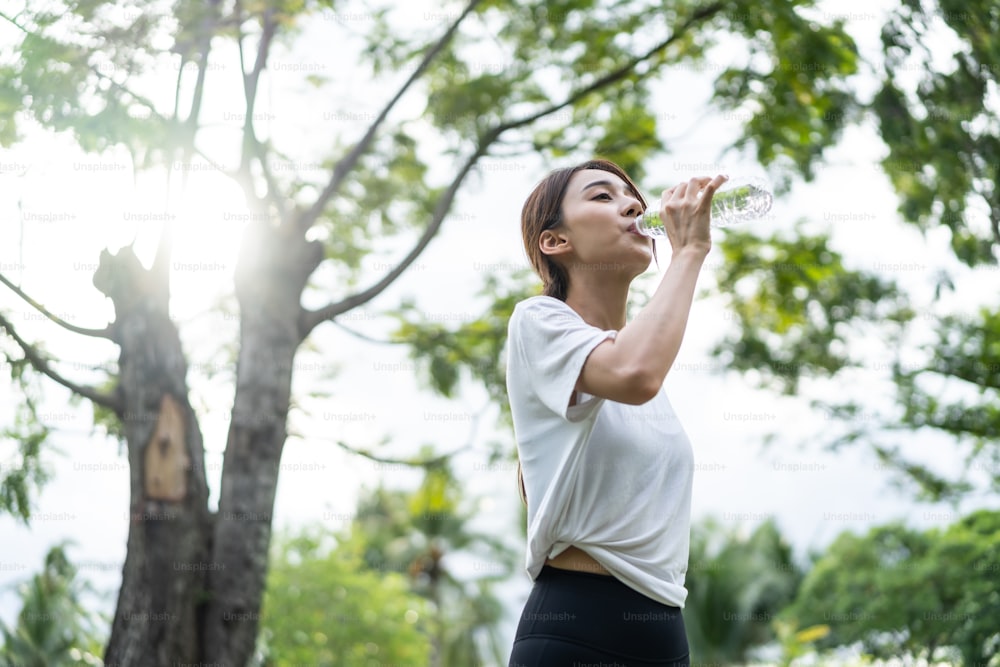 L'atleta asiatico beve una bottiglia d'acqua dopo l'esercizio nel parco. La bella sportiva attraente attiva si prende una pausa dopo lo yoga, l'allenamento in esecuzione all'aperto la sera per l'assistenza sanitaria in giardino.