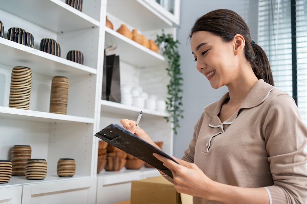 Asian beautiful woman check vase goods order for customer from shelf. Young attractive business girl working to preparing parcel boxes checking ecommerce shipping online retail to sell at home store.