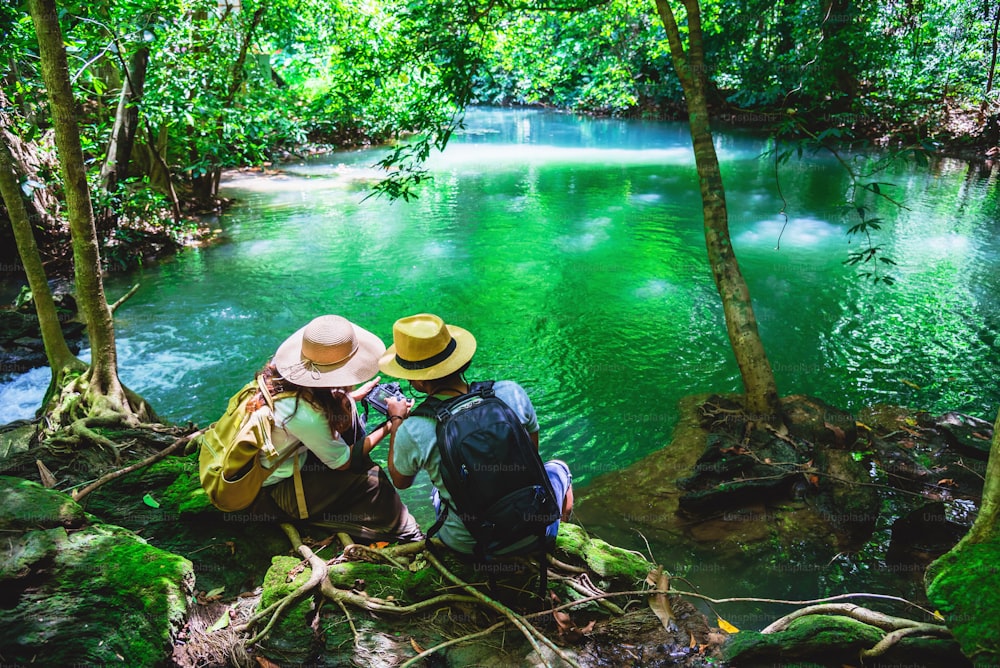 旅行者、バックパックを背負ったカップル、岩に座ってリラックスしてください。緑のジャングルで自然を旅し、滝の景色を楽しんでいます。観光、ハイキング、自然研究。旅行中のカップル、休日に自然の写真を撮る。エメラルドストリーム。