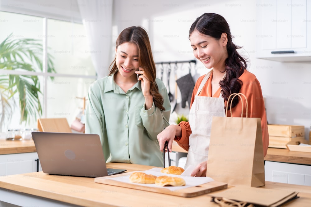 Asian beautiful business woman and friend bake bakery and sale in house. Attractive lesbian couple feel happy and exciting for success in entrepreneur sell food order from home then high five together