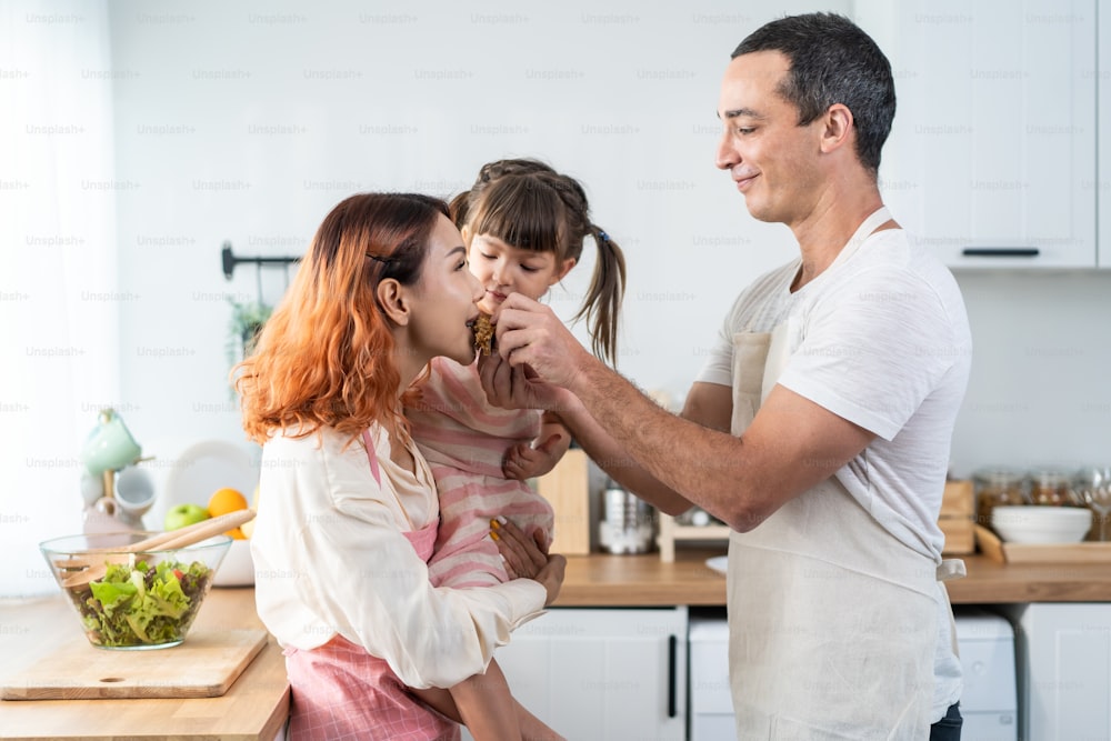 Asian happy family spend free leisure time in kitchen at home together. Loving husband feed cupcake to his beautiful wife while holding young kid daughter, prepare to cook foods for breakfast in house