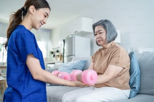 Asiatische behinderte alte Frau, die Physiotherapeutin mit Unterstützung der Krankenschwester macht. Ältere �ältere behinderte Frauen sitzen auf dem Sofa im Wohnzimmer mit Hanteln Trainingsübung für Patienten in der häuslichen Pflege.